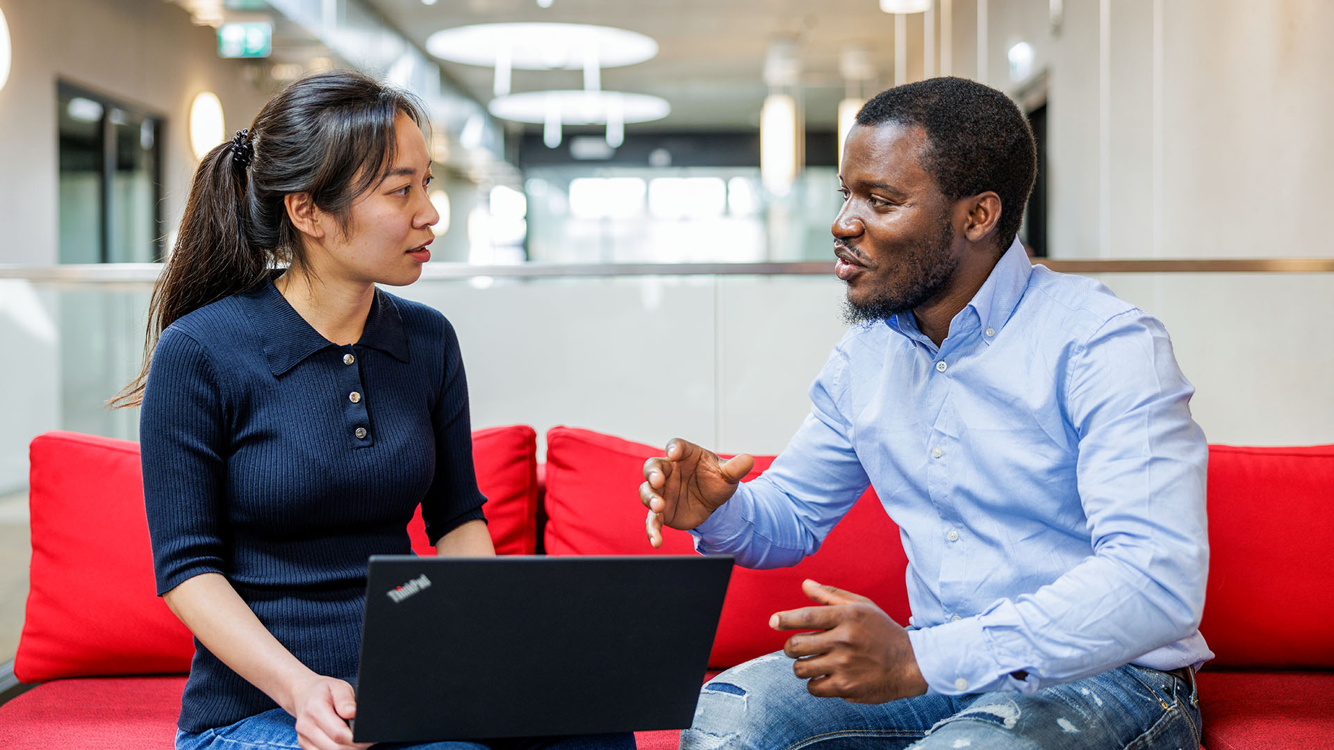 Two ASML employees engaging in a discussion while sitting on a red couch in a modern office space, with one person holding a laptop. Two ASML employees engaging in a discussion while sitting on a red couch in a modern office space, with one person holding a laptop.