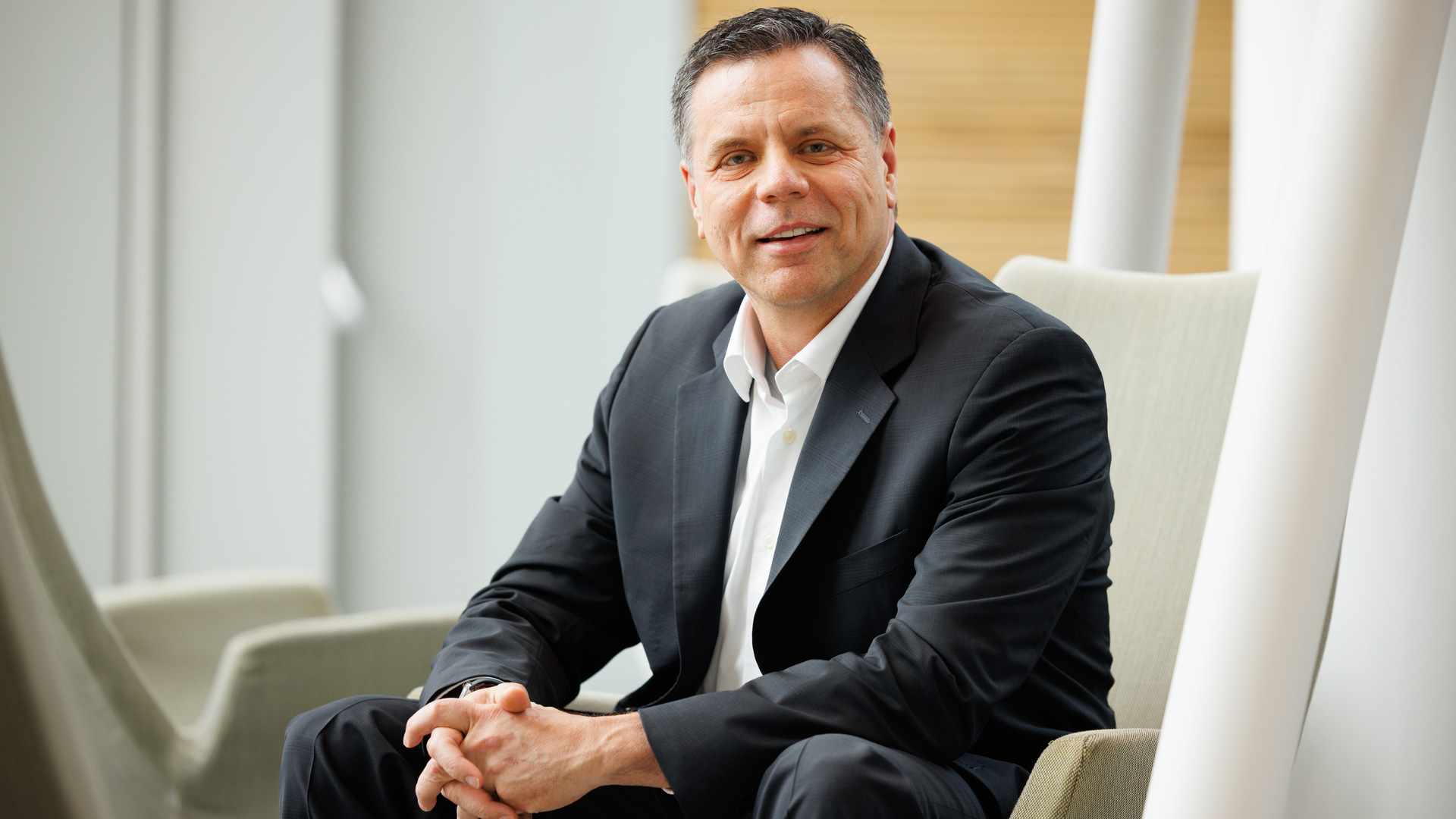 A man with gray hair, wearing a black suit and white shirt with no tie, leans forward in his chair and smiles at the camera. 