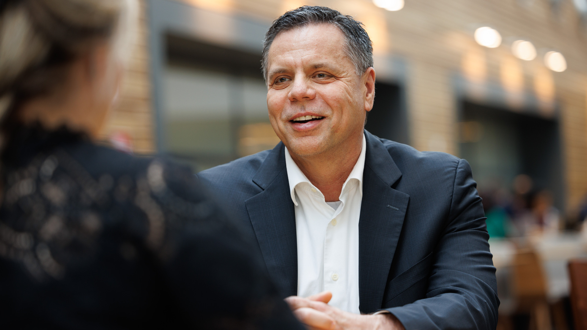A man wearing a dark blue suit and white shirt with no tie sits at a table in a cafeteria and smiles at his interviewer.