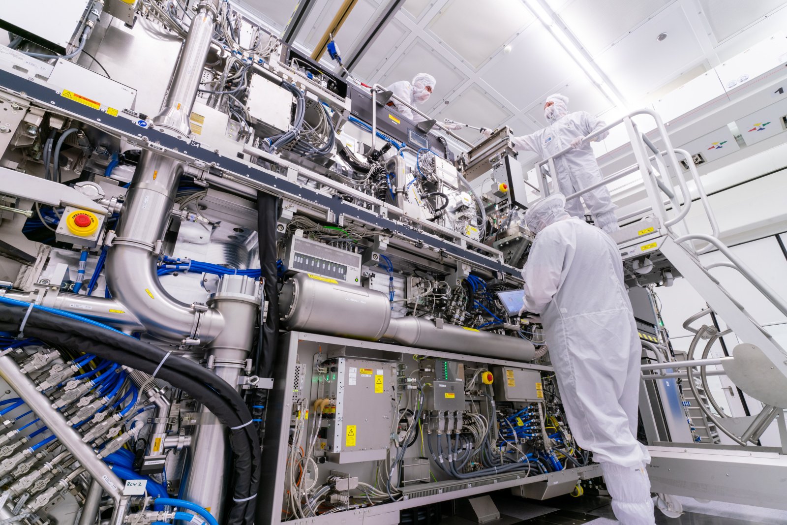 a low-angle photo of three people in cleanroom suits working on a huge lithography system with many wires and pipes. 