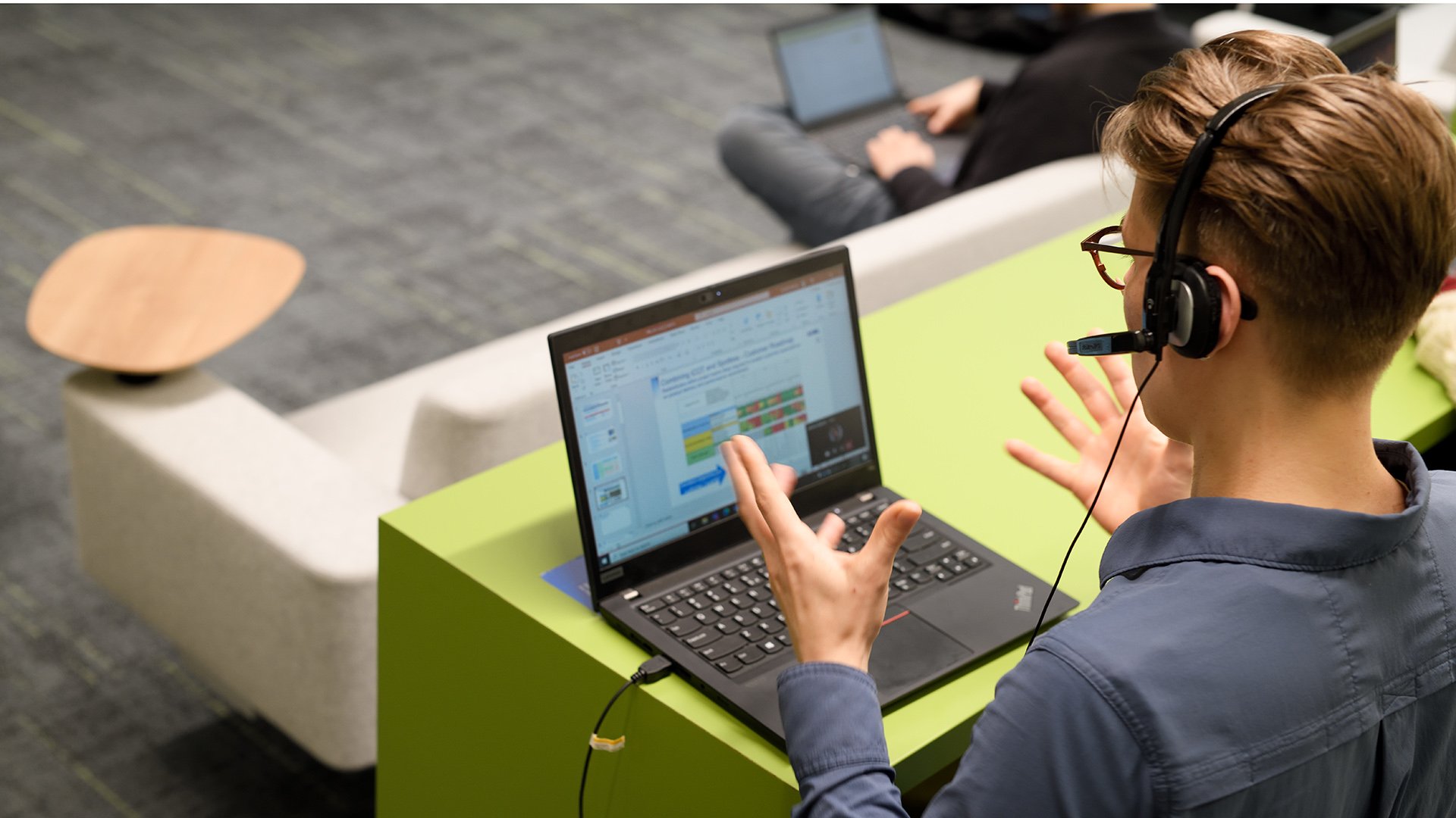 A young person wearing headphones gestures with hands while sitting in front of a laptop.