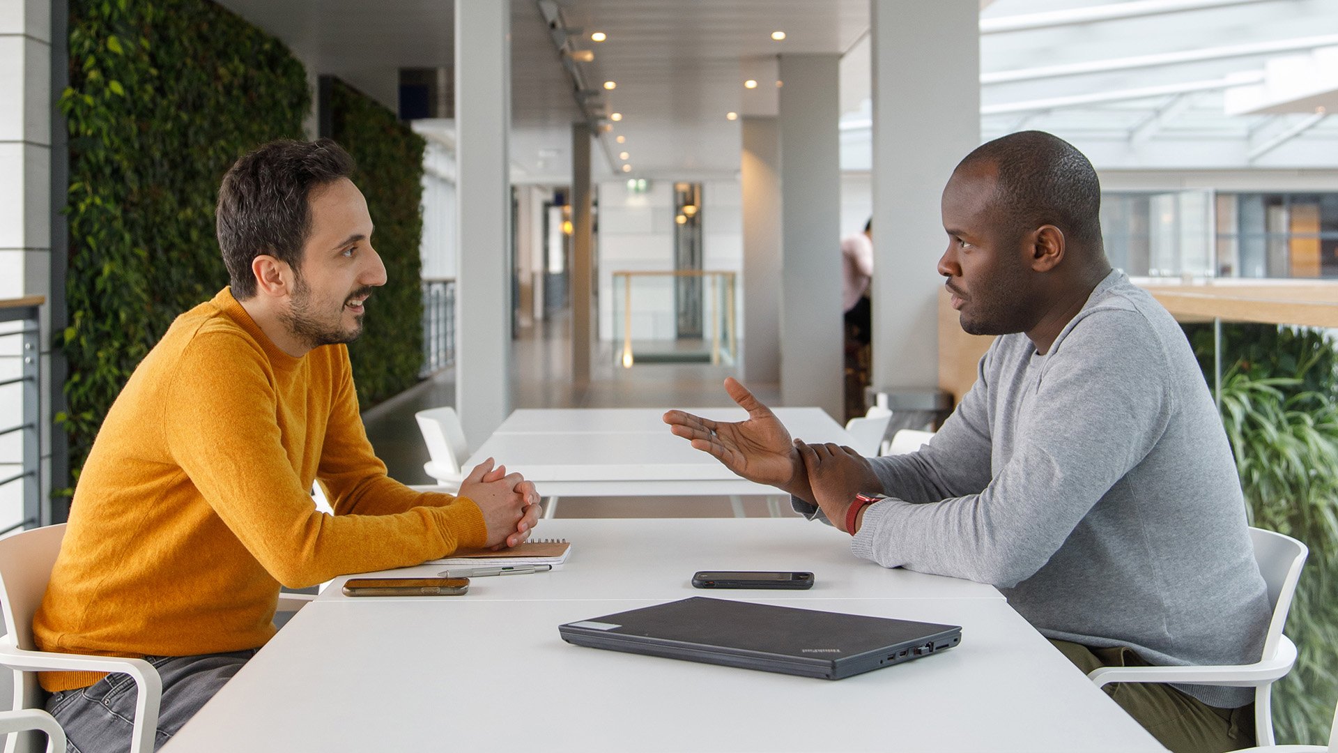 Two ASML colleagues engaging in a conversation at a white table, with one person explaining something with his hands, and a closed laptop, suggesting a collaborative work session. Two ASML colleagues engaging in a conversation at a white table, with one person explaining something with his hands, and a closed laptop, suggesting a collaborative work session.