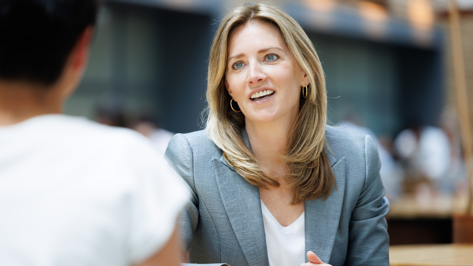 A blonde woman in a gray blazer and white shirt sits at a table in a cafeteria and smiles at her interviewer.