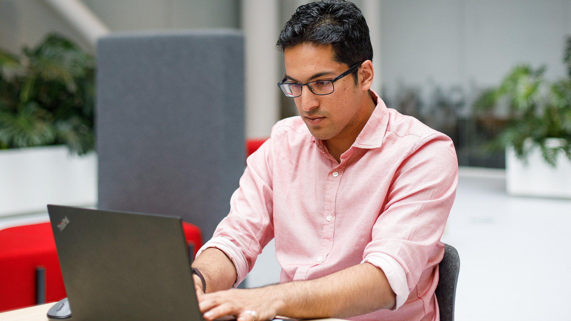 An ASML employee wearing glasses and a pink shirt sitting at a desk and working on a laptop. An ASML employee wearing glasses and a pink shirt sitting at a desk and working on a laptop.