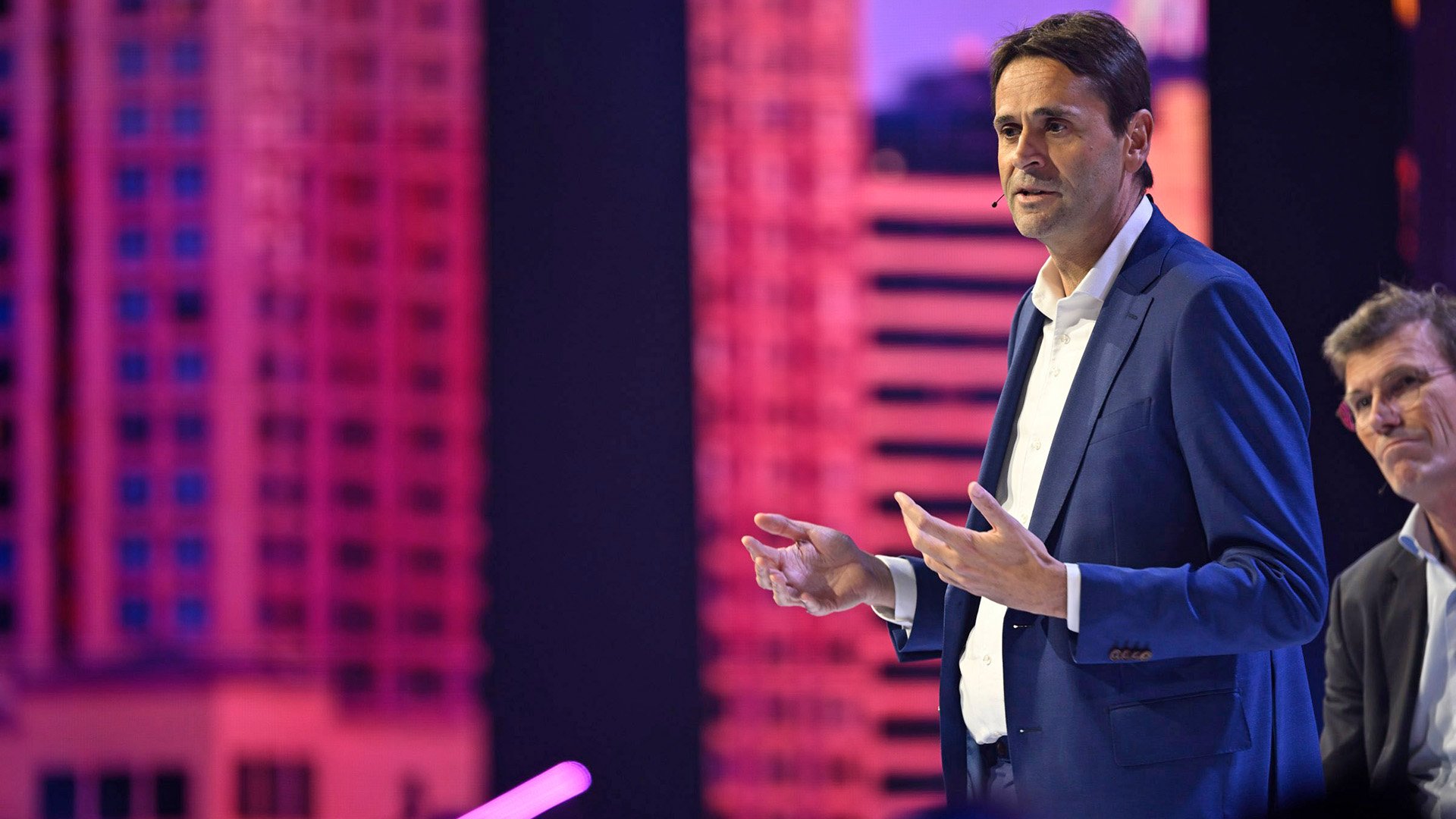 Christophe Fouquet, dressed in a navy blue suit and white shirt, gesturing with open hands against a vibrant pink digital backdrop, communicating a point during a presentation. Christophe Fouquet, dressed in a navy blue suit and white shirt, gesturing with open hands against a vibrant pink digital backdrop, communicating a point during a presentation.