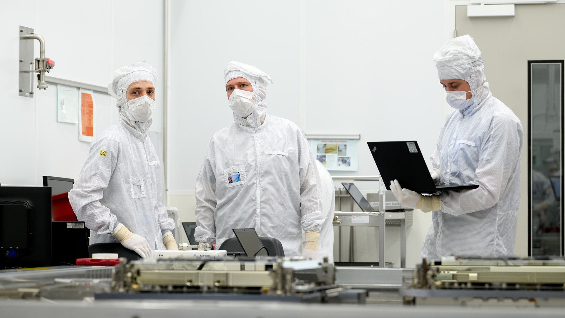 One person working on a laptop while two other people look across the room One person working on a laptop while two other people look across the room