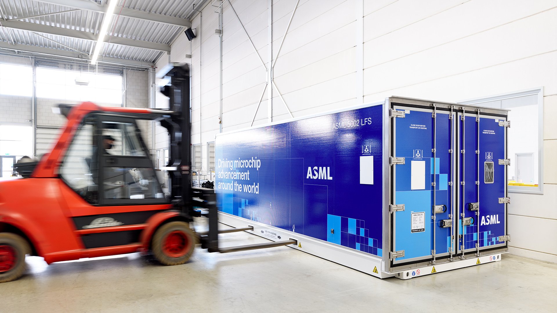 Inside a warehouse, a red forklift moves towards an ASML shipping container. Inside a warehouse, a red forklift moves towards an ASML shipping container.