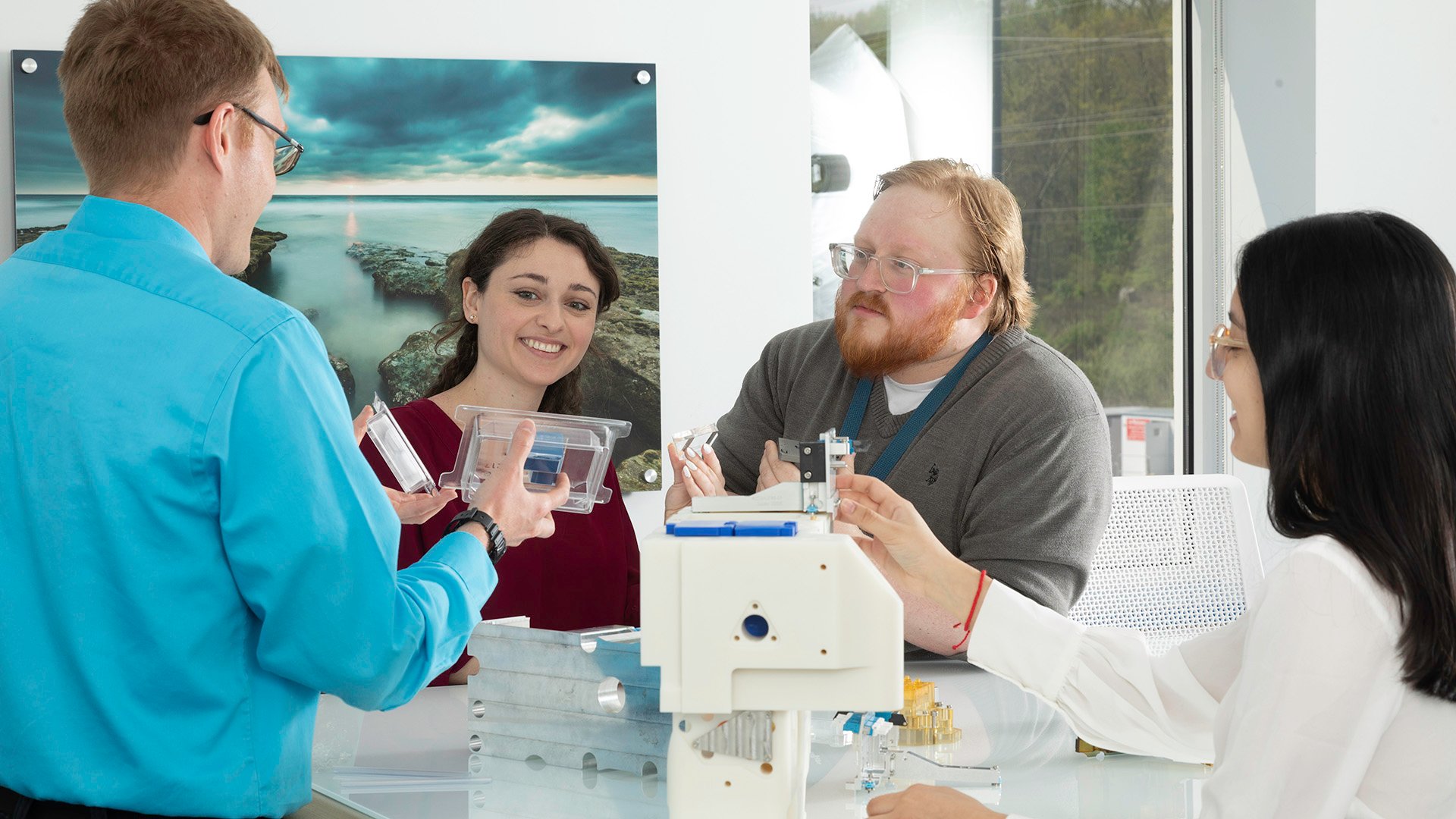 A group of two men and two women smile as they gather around a lab table where one of the men holds a technical instrument.